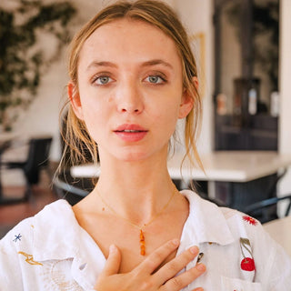 Young woman wearing carnelian necklace featuring 5 carnelian stones stacked vertically hanging from a delicate gold chain. She looks into the camera and touches her hand to her chest.