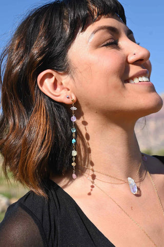 Woman wearing long drop earrings featuring different chakra crystals while standing in the sun and smiling.